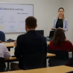 A professional classroom scene showing a diverse group of students and a teacher engaged in an educational activity, with a visible ICDL certification poster on the wall, modern desks, computers, and a whiteboard displaying collaborative learning symbols, all in a clean, realistic style